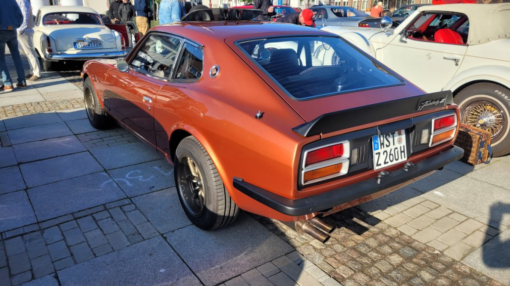 Auf einer Autoshow im Freien parkt zwischen anderen Oldtimern ein orangefarbener Oldtimer-Sportwagen.